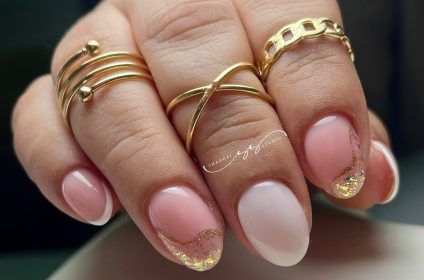Manicura de estilo francés moderno con ondas de brillo dorado sobre base rosa pálido y anillos de oro, por Shaddai Studio.